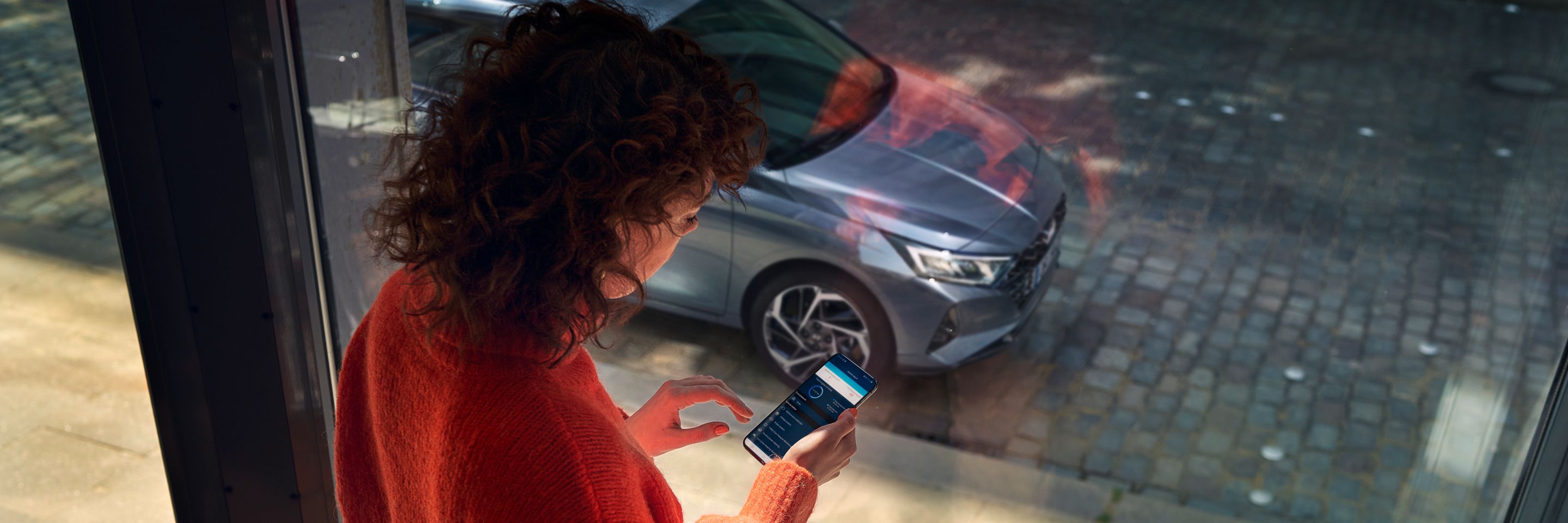 Žena dívající se na aplikaci Hyundai Bluelink na svém smartphonu, její Hyundai i20 je zaparkovaný venku.